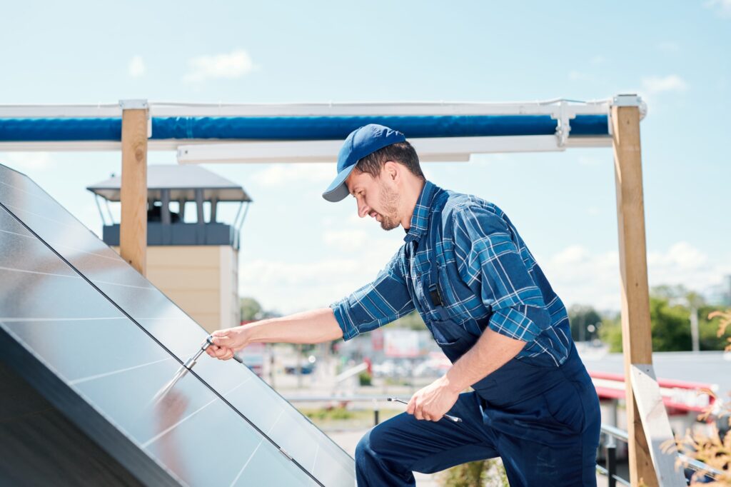 Young technician master in workwear bending over solar panel on the roof